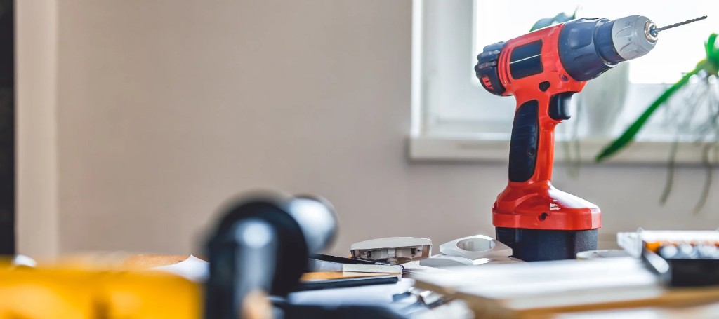 A red-and-blue cordless power drill stands upright on a workshop table beside tools, with a window and plant in the background, suggesting a practical DIY workspace.