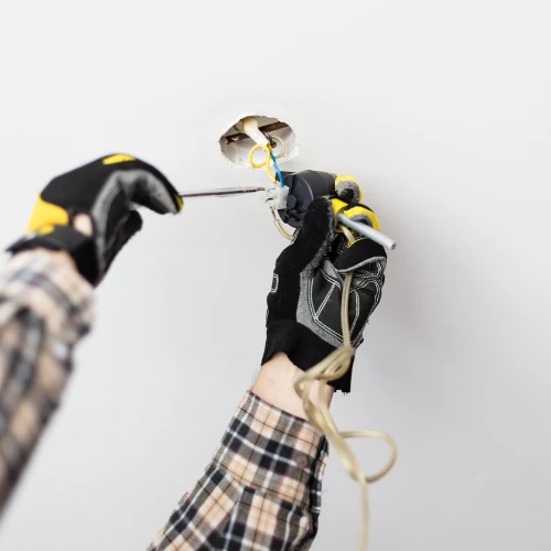 A worker in black gloves uses pliers to handle exposed wiring in a wall opening, focusing carefully on an electrical repair in progress.