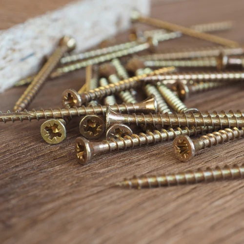 A close-up of long brass-colored wood screws scattered across a dark wooden surface, with a blurred cloth in the background; sturdy, workshop-ready feel.