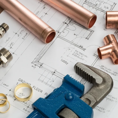 Copper pipes, fittings, a blue pipe wrench, and measuring tape lie on an architectural floor plan, suggesting plumbing work in progress.
