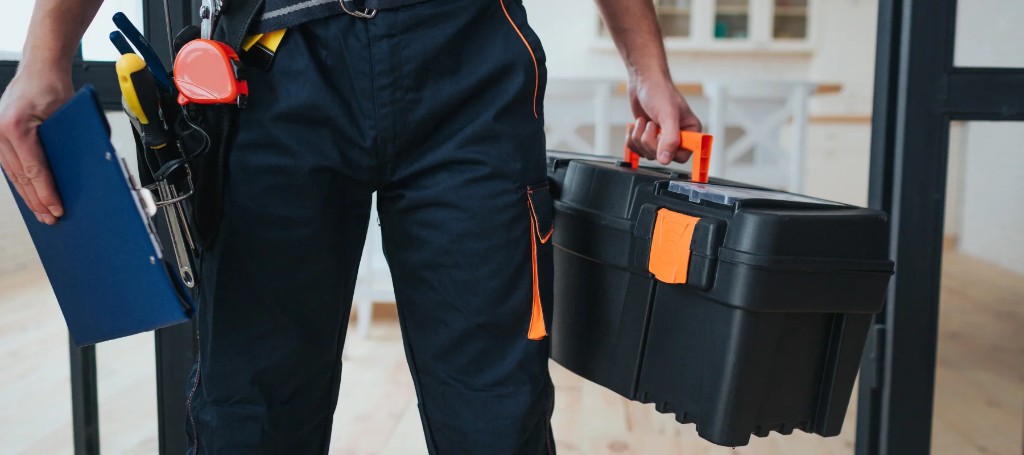 A tradesperson in black workwear carries a large toolbox and blue folder, with tools on a utility belt in a bright indoor workspace.