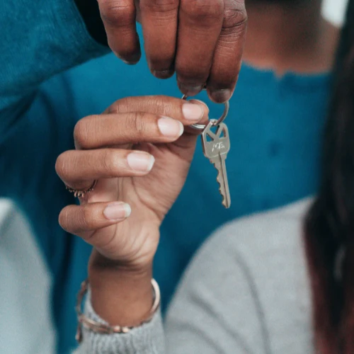 A person in a gray sweater reaches for a silver key offered by another hand, capturing a quiet, hopeful moment of receiving access to a home.